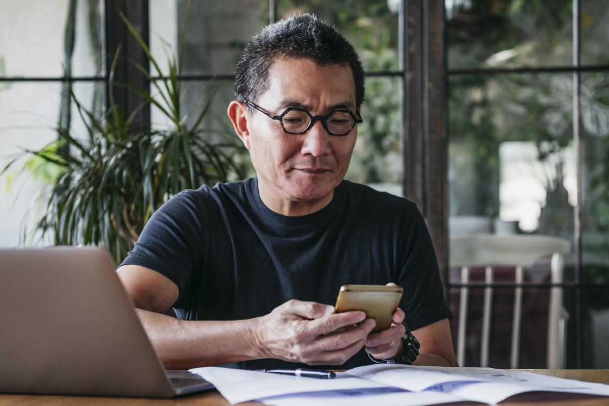 mature man texting on cell phone with paperwork and laptop