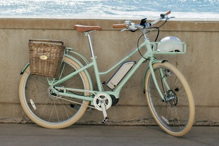 A light pastel green e-bike with a basket at the back sitting against a concrete wall by the ocean.