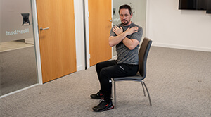 Andrew Scard demonstrates seated trunk rotation on a chair with his arms crossed on his chest as he turns to his left