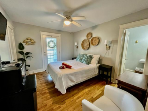 Interior of a room at the Carolina Beach Inn featuring a comfortable bed with white linens, decorative pillows, and wall art. The room has a ceiling fan, hardwood floors, and an en-suite bathroom.