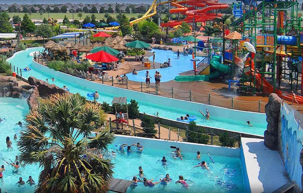 A busy water park with various pools, slides, and colorful umbrellas. Many people are enjoying swimming and leisure activities. Trees and greenery surround the area.