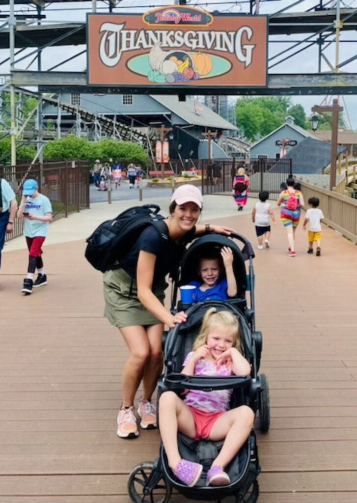 An adult and two children in a stroller pose for a photo on a wooden walkway under a sign that reads 