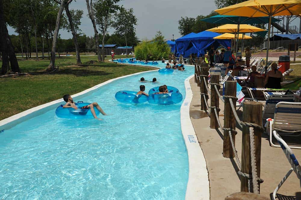 Several people float on blue inner tubes along a lazy river at an outdoor water park. Trees and umbrellas provide shade, while lounge chairs and tables are set up along the side.