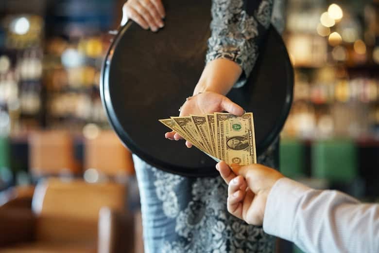 A person hands several US dollar bills to another individual holding a black serving tray in a restaurant setting.