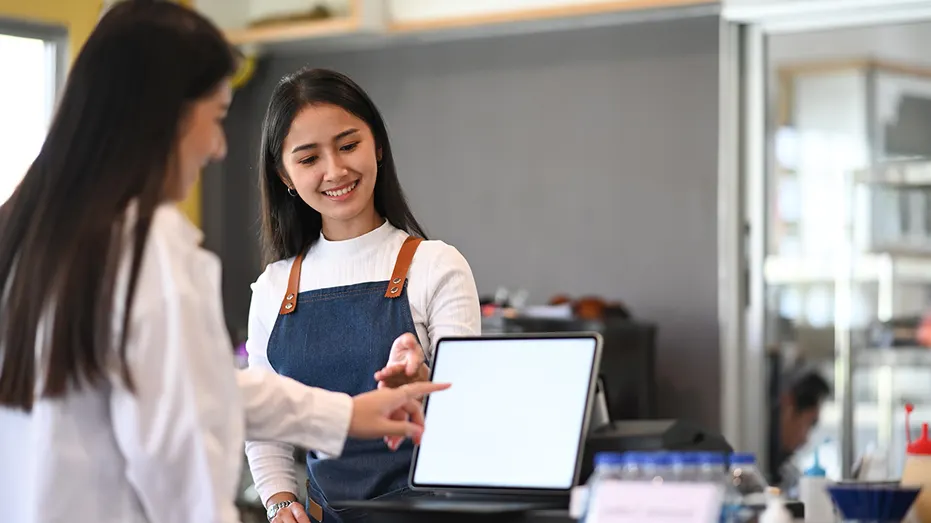 Two women are at a counter. One woman, wearing an apron, is pointing at a screen while the other woman looks and points as well. The background includes shelves with various items.