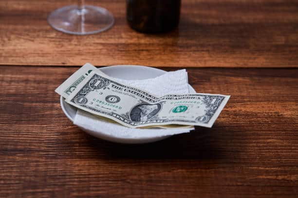 Two one-dollar bills are placed on a white napkin inside a bowl on a wooden table, next to a partially visible wine glass.