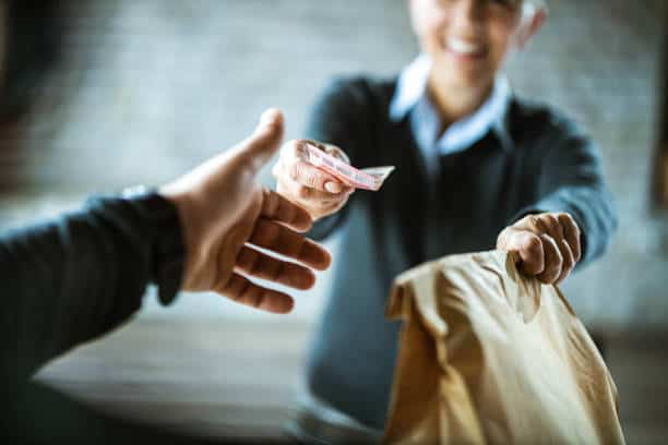 A person hands over money while receiving a brown paper bag from another person.