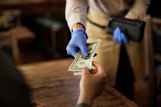 A person wearing blue gloves hands over money to another individual across a wooden table.