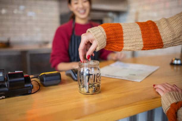 Person placing coins into a clear tip jar on a wooden counter, with a smiling barista in the background.