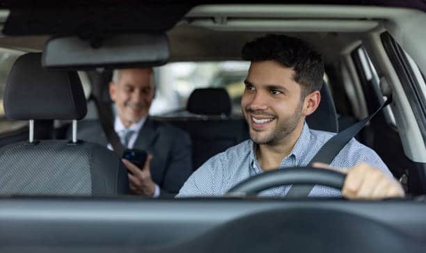 A man drives a car with a smiling passenger in the back seat holding a smartphone.