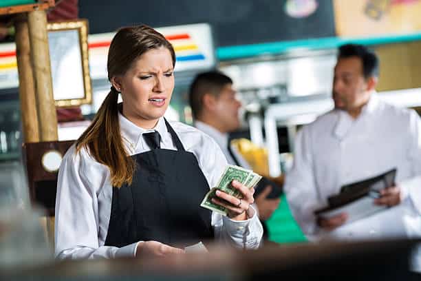 A waitress in a black apron is counting money and appears concerned. Two chefs are in the background, one holding a menu. The setting seems to be a busy restaurant.