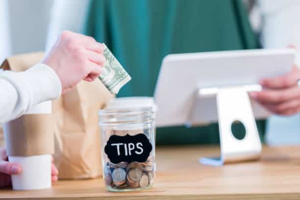 A hand places a dollar bill into a glass jar labeled 