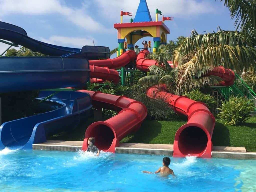 Two people emerge from red water slides into a pool, with a colorful water playground structure in the background. Don't forget to check for Legoland California coupons to make your visit even more enjoyable!