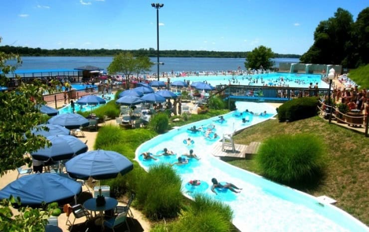 A busy outdoor waterpark with a lazy river, pools, lounge chairs, umbrellas, and people swimming and relaxing. The park is near a large body of water with trees in the background.