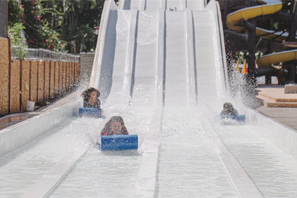 Four people slide down parallel water slides, splashing into the shallow water at the bottom.