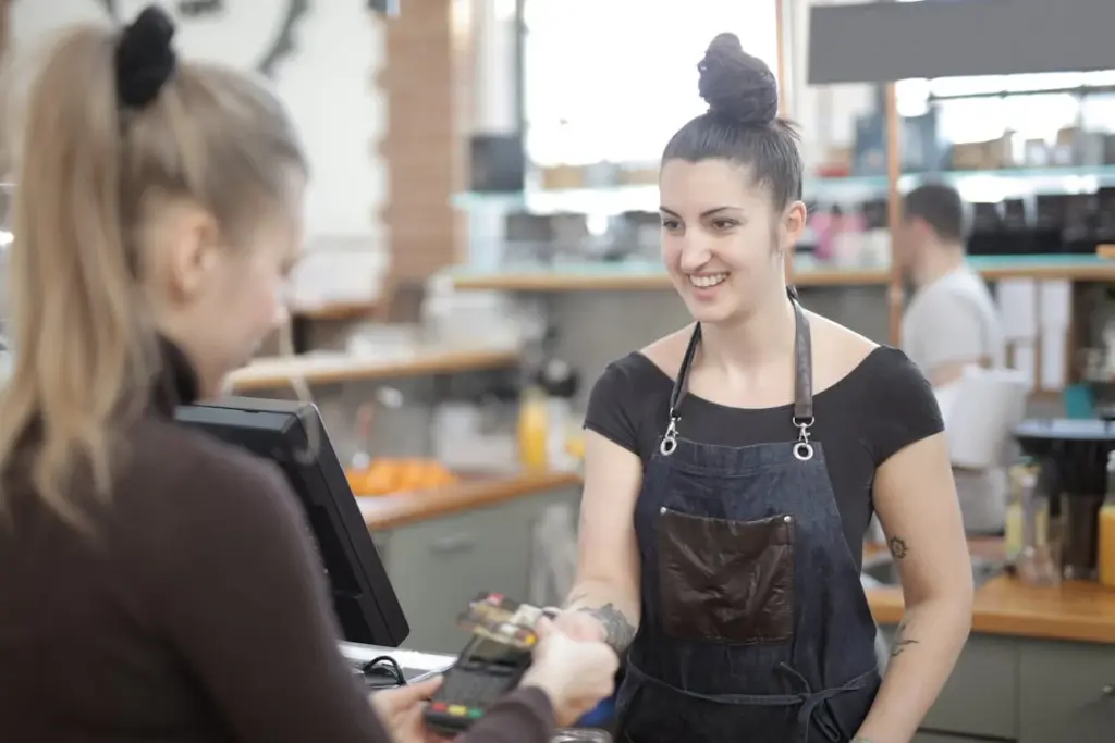 A cashier in a dark apron smiles while handing a payment terminal to a customer at a retail counter. The customer, reflecting how Americans are tired of tipping, holds their card over the terminal.