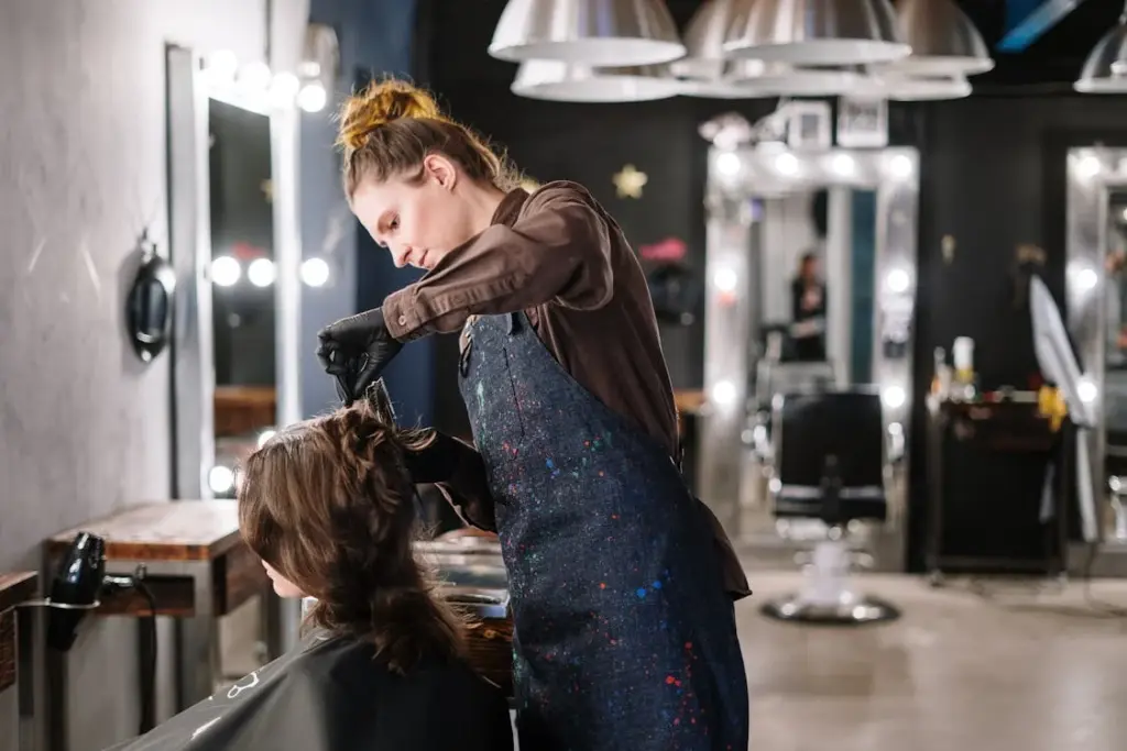 A hairdresser wearing gloves and an apron styles a client's hair in a modern salon with mirrors and hanging lights.