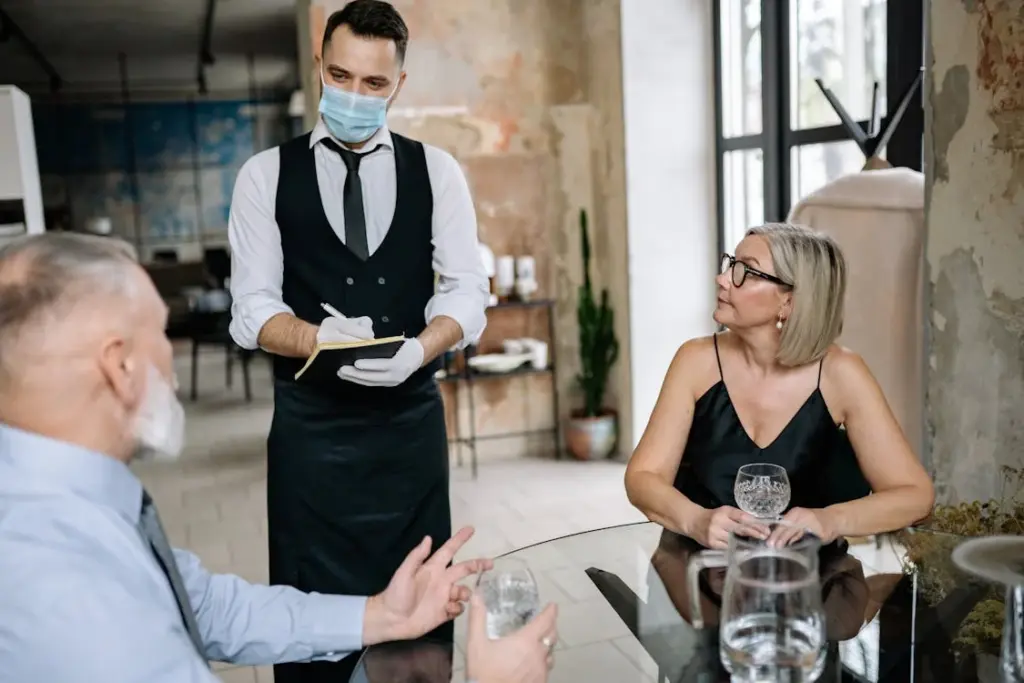 A waiter, wearing a mask and gloves, takes an order from a seated man and woman in a restaurant with a rustic interior. The establishment prides itself on gratuity-free service, embracing a fancy way to say no tips by including all charges in the bill upfront.