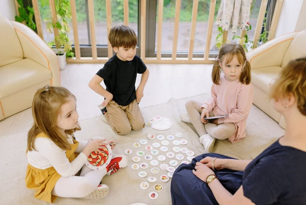 Three children sit on the floor, playing a game with circular cards while an adult watches. The setting appears to be a bright, cozy living room with beige furniture and plants near the window.