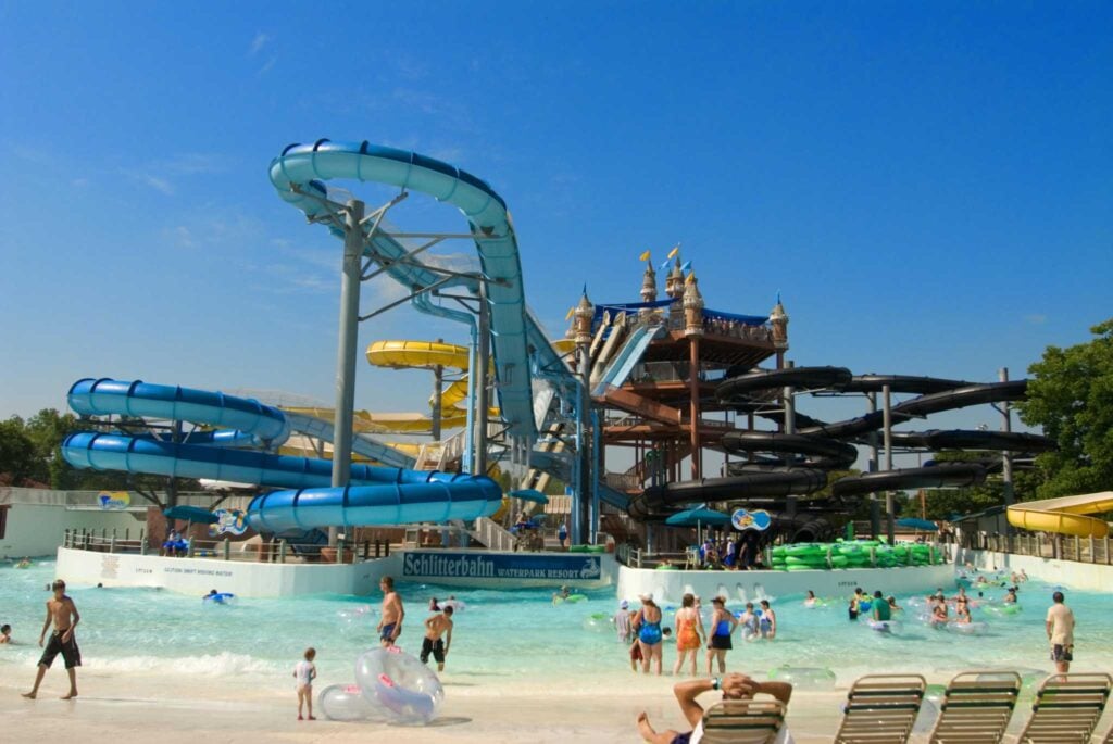 A crowded water park features multiple winding water slides and a shallow wave pool filled with people. Lounge chairs line the foreground as visitors enjoy the sunny day.