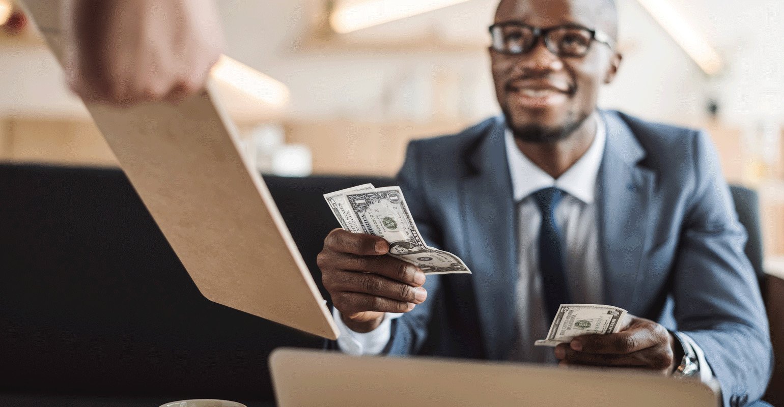 Smiling man in a suit seated at a desk handing cash to a person holding a folder.