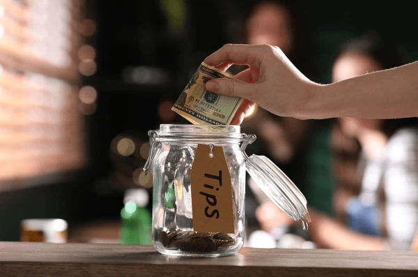 A hand places a folded dollar bill into a clear glass jar labeled 