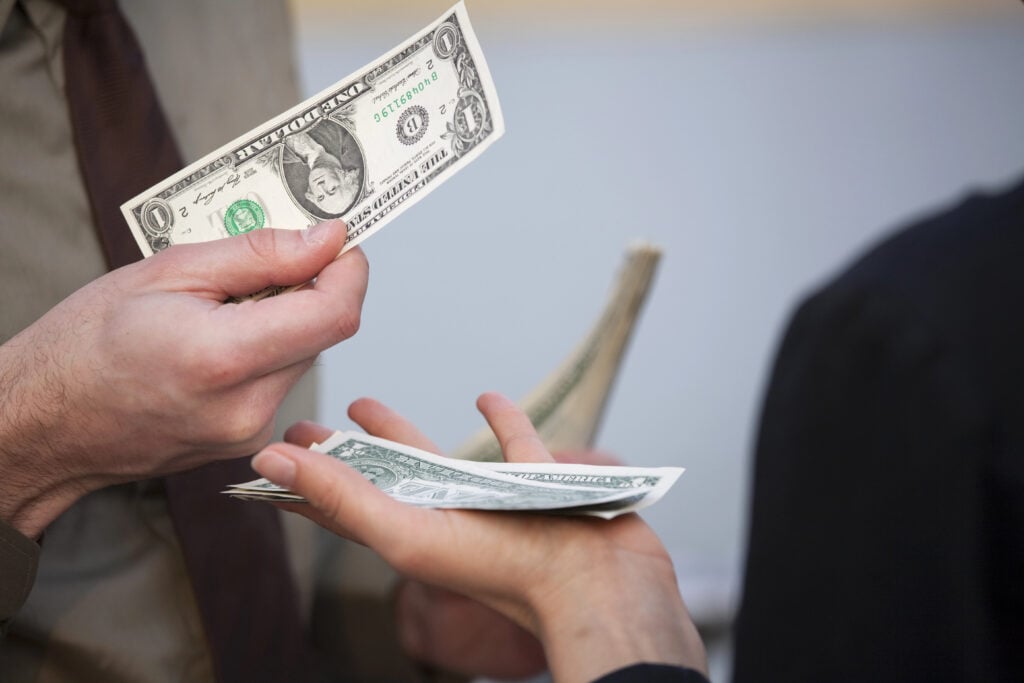 A person hands over a one-dollar bill to another person, who is holding multiple dollar bills, as they discuss things people pay for.