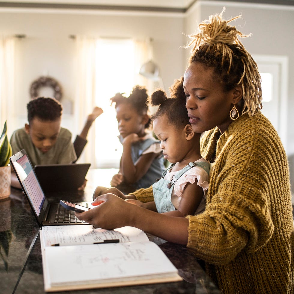 mother working from home while holding toddler, family in background