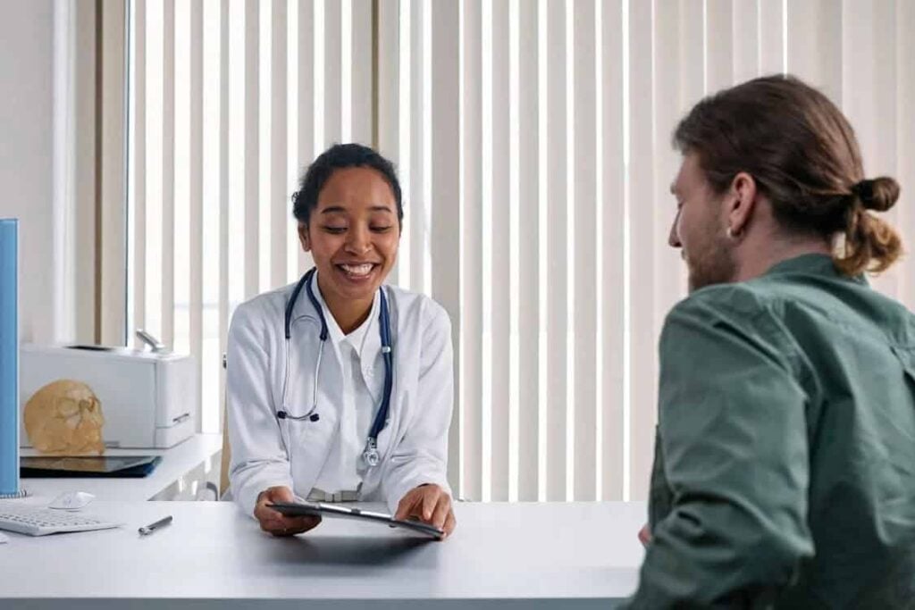 A doctor wearing a white coat and stethoscope shows a tablet to a seated patient in an office.