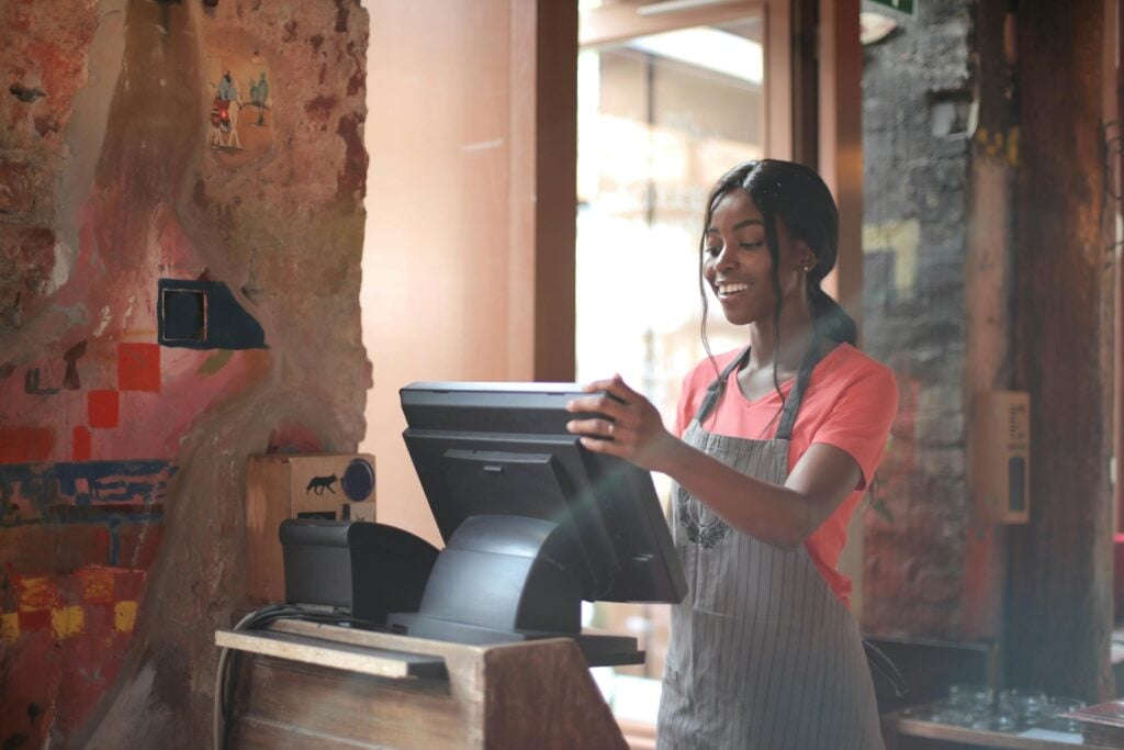 A woman wearing an apron stands at a cash register with her hands on the screen, smiling in a warmly lit setting with colorful wall decor.