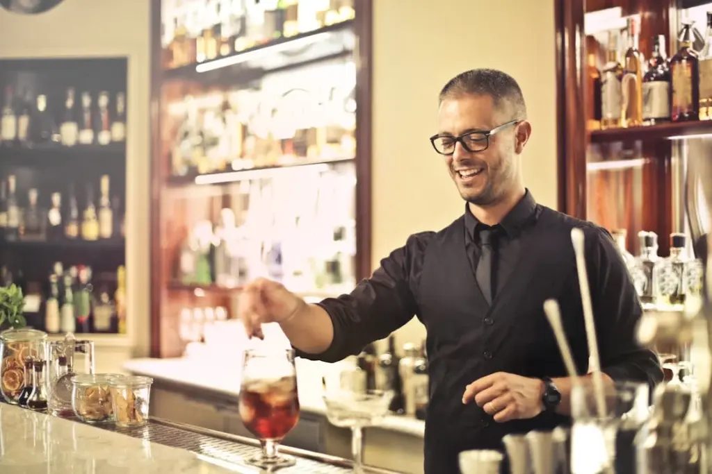 A bartender with glasses and a dark vest stirs a drink at a well-stocked bar.