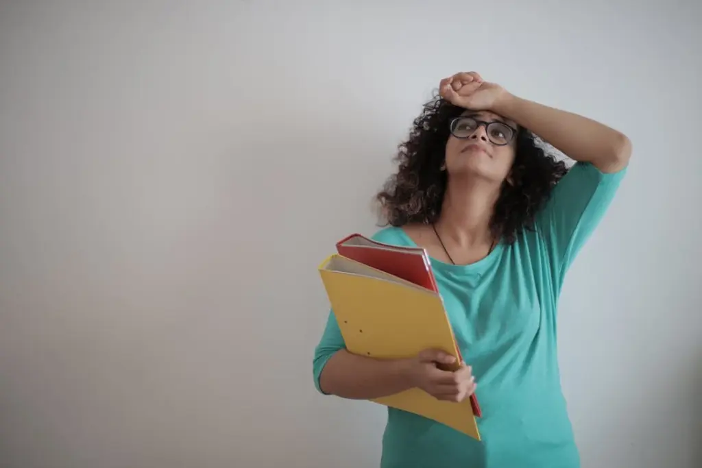 A working-class individual with curly hair and glasses, wearing a green shirt, holds folders and rests their head on their hand against a white background.