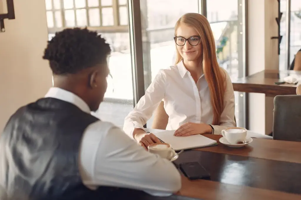 A woman and a man sit across from each other at a table in a cafe, both with coffee cups. The woman has a notebook, and they appear to be engaged in a discussion.