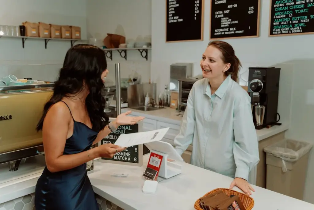 A woman in a blue dress is talking to a barista behind the counter at a cafe. The barista is wearing a light blue shirt and smiling. There are menu boards and coffee equipment in the background.