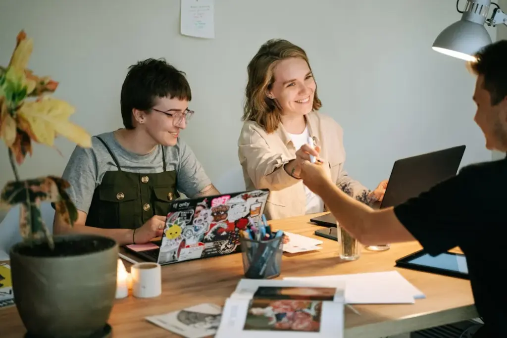 Three people collaborate at a desk, two with laptops and one handing over papers. The workspace is scattered with documents, a plant, and a lit candle. They appear engaged in a discussion.