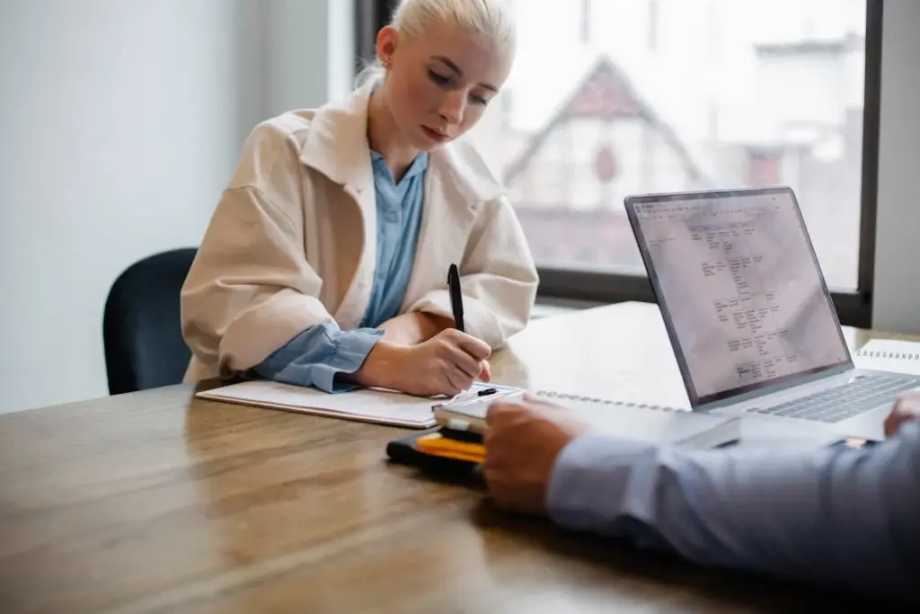 A person with blonde hair writing on paper at a desk, while another person sits across with an open laptop.