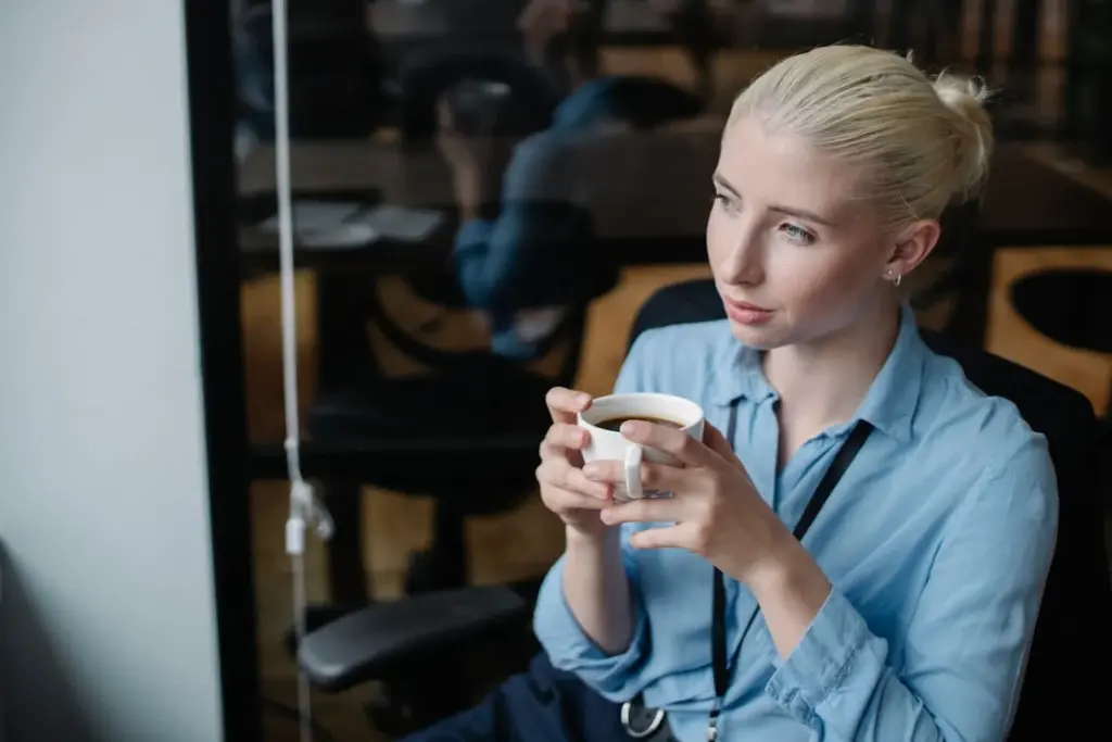 A person with light hair in a bun sits in a chair, holding a white coffee cup, looking to the side thoughtfully. They are wearing a blue shirt and a lanyard.