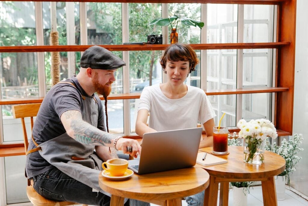 Two people sitting at a table in a café, engaging with a laptop. A drink and flowers are on the table. The café has large windows and wooden furniture.