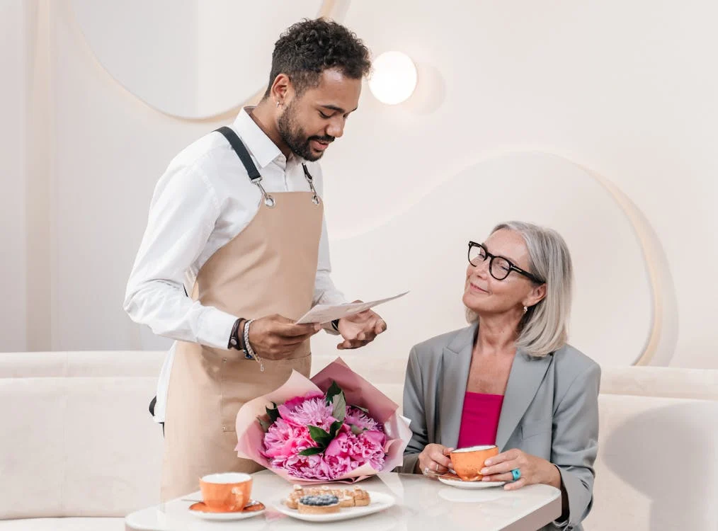 A waiter in an apron serves a seated woman holding a cup. A bouquet of pink flowers and a pastry are on the table. The establishment prides itself on elegance, even displaying a fancy way to say no tips on credit card at each table.