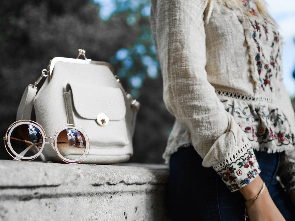 woman wearing beige and red floral top leaning on gray concrete slab with white leather bag ontop