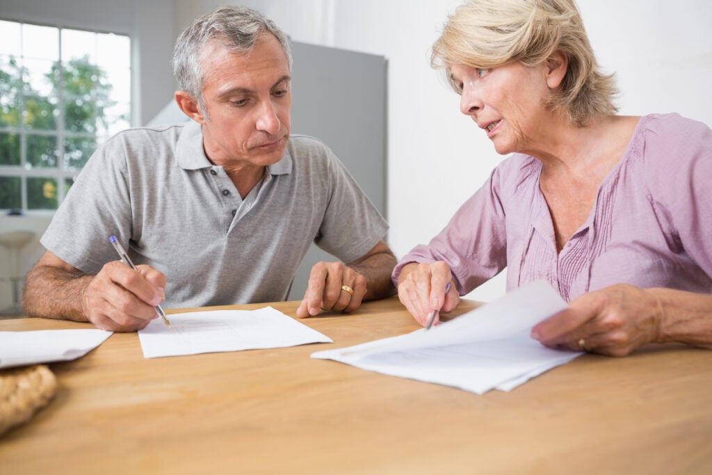 Two older adults, a man and a woman, sit at a table reviewing documents together in a well-lit room. They appear focused and engaged in their discussion about what to include in their will.