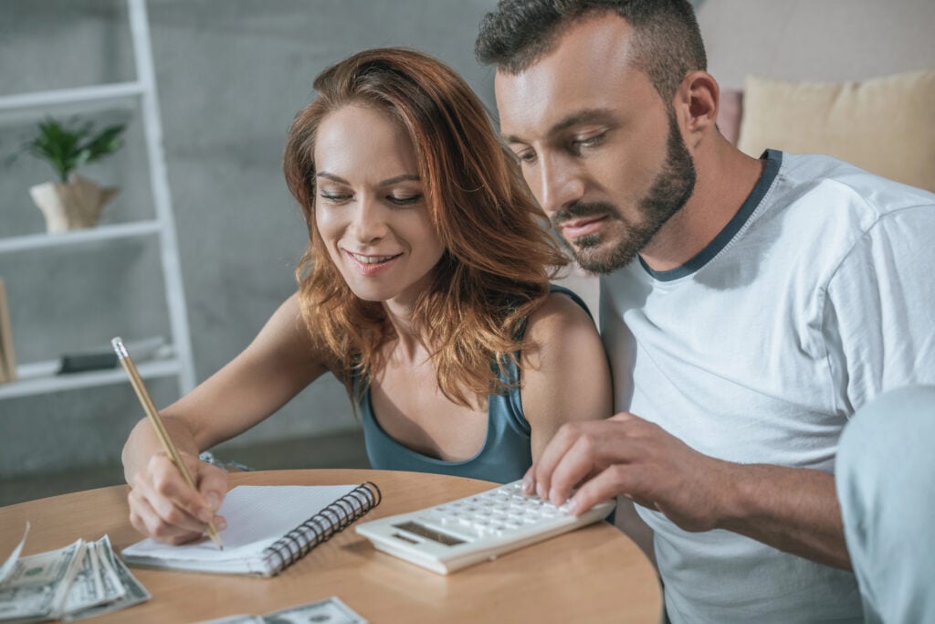 Two people sit at a table, one writing in a notebook with a pencil, the other using a calculator. Cash is spread out on the table beside them. They appear focused on their task, perhaps figuring out how to save on monthly bills.