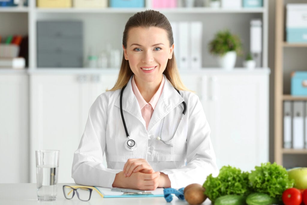 A female doctor with a stethoscope sits at a desk surrounded by fresh vegetables in a medical office.