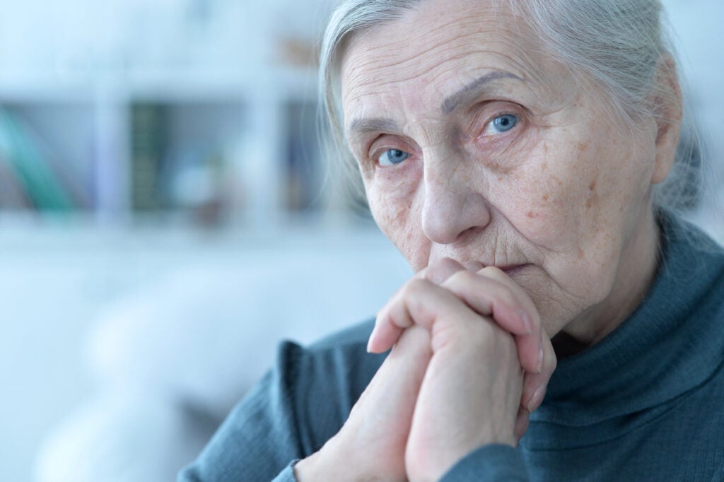 An elderly woman with gray hair and a blue sweater looks pensively at the camera, resting her hands against her chin in a brightly lit room.