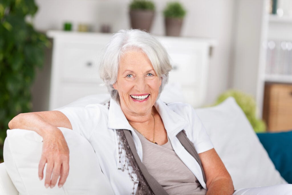 Elderly woman with short white hair, wearing a white shirt and gray scarf, sitting on a couch and smiling.