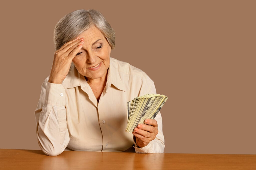 A pensive elderly woman, sitting at a table, holds a stack of money in her hand while resting her other hand on her forehead.