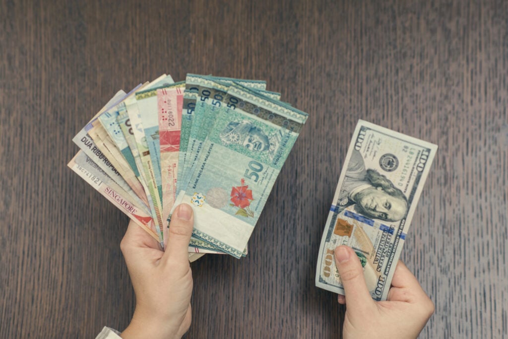 Person holding a fan of various foreign currencies in the left hand and U.S. dollars in the right hand over a wooden table.