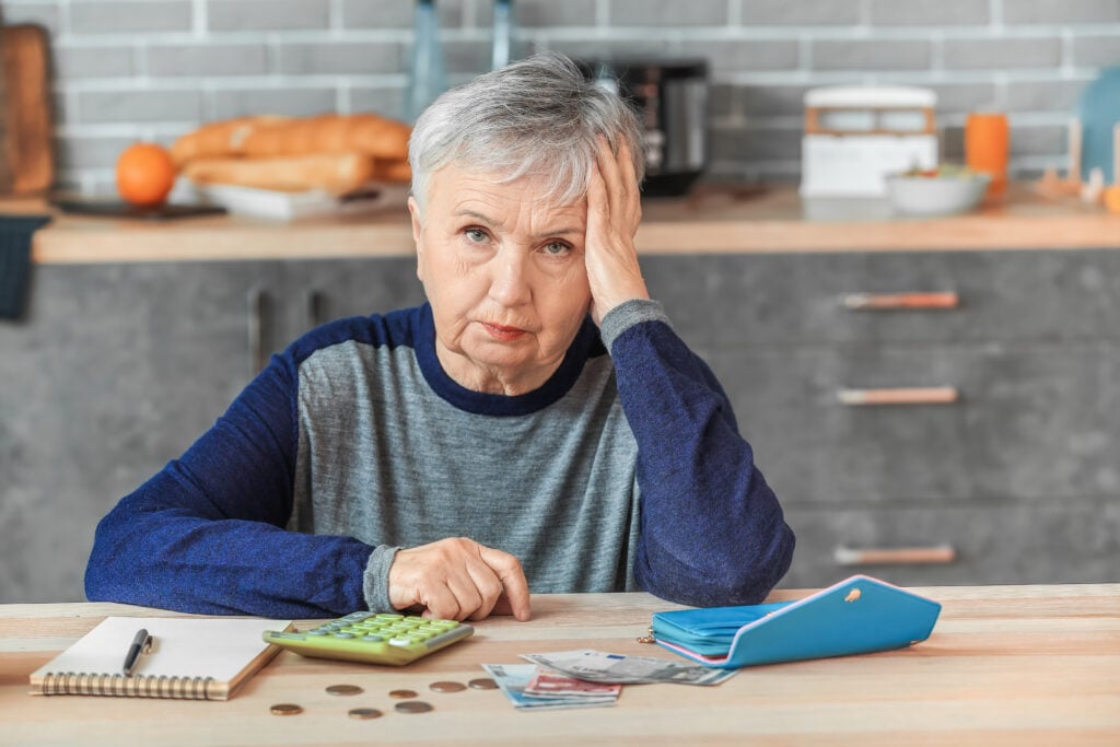 An elderly woman sitting at a kitchen table, looking concerned, with a calculator, notebook, coins, and bills spread out in front of her.