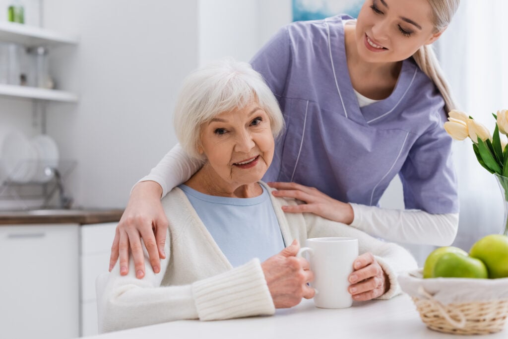 Elderly woman in a light blue shirt holding a white mug, seated at a table with a young caregiver in a purple uniform standing beside her and smiling, discussing things to stop doing if 75.