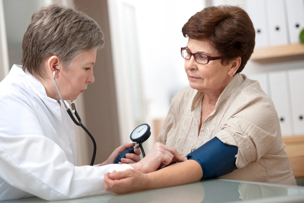 A healthcare provider measures the blood pressure of an older adult woman using a sphygmomanometer in a clinical setting, reminding her to stop doing these if 75: strenuous exercises and unapproved medications.